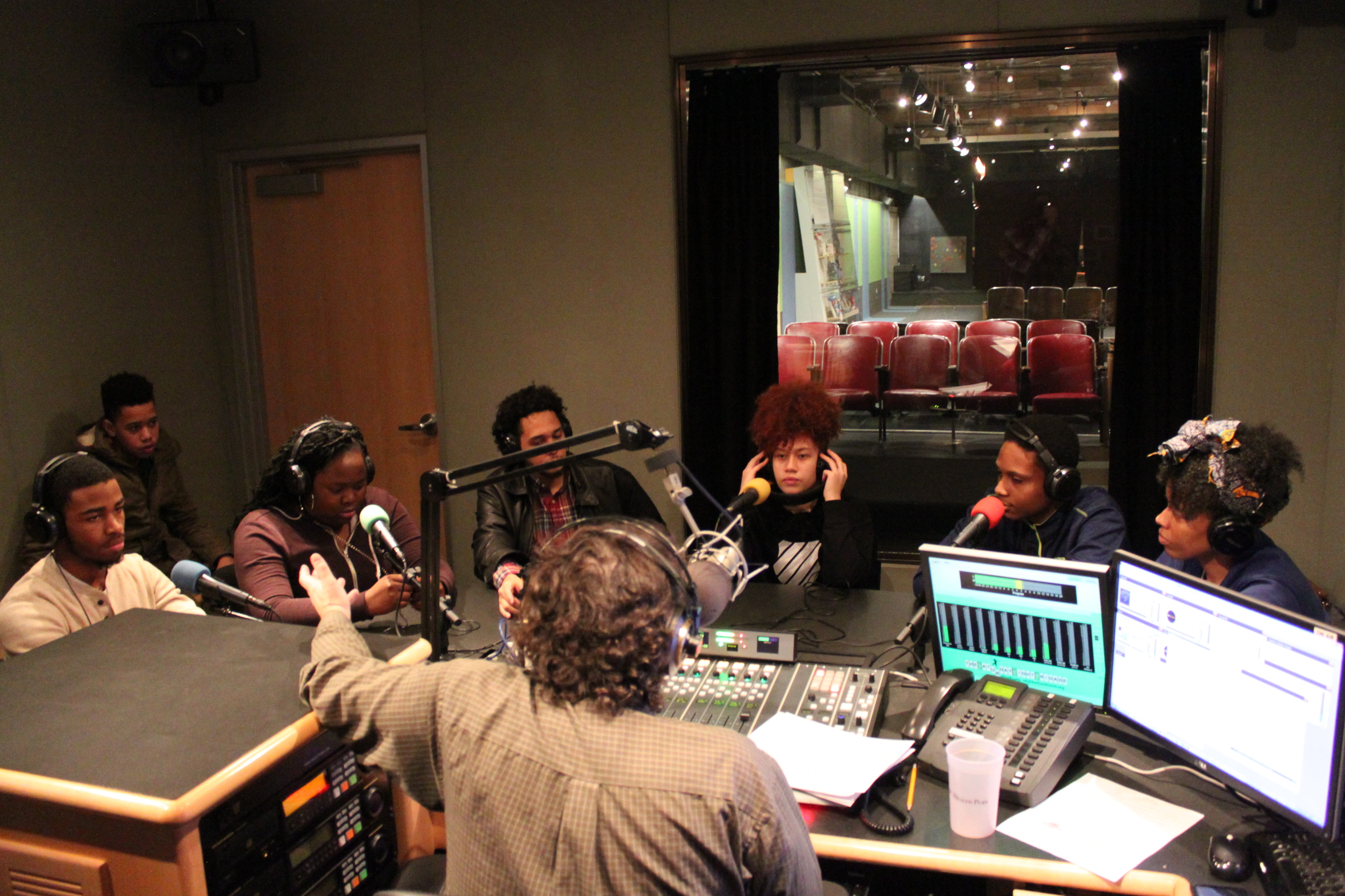 Six teens wearing headphones sitting behind microphones and one youth sitting in the back corner being interviewed by an adult wearing headphones speaking into a microphone sitting behind a mixing board and gesturing with their hand in a radio studio