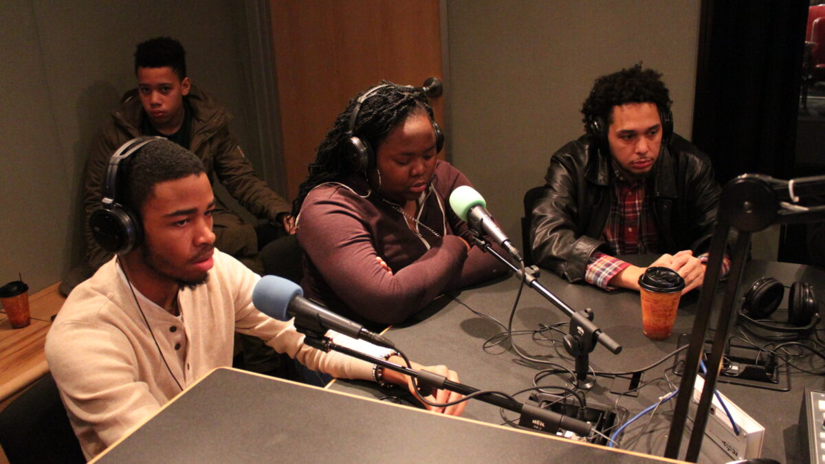 Three teens wearing headphones sitting behind microphones and one teen sitting in the back corner in a radio studio