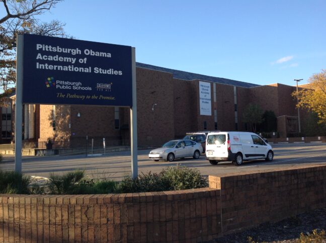 A large brick building with three cars parked in the parking lot in front, along with a dark blue sign with the text "Pittsburgh Obama Academy of International Studies" in white, along with a multicolored square logo and the text "Pittsburgh Public Schools" in white, a graphic of the text "Excellence For All" in white, with a yellow star shooting behind "Excellence", and the text "The Pathway to the Promise" in yellow