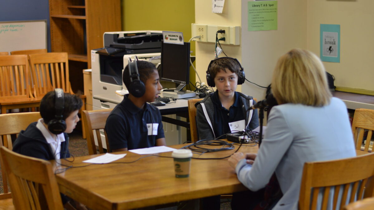 Three youth wearing headphones interviewing an adult speaking into a microphone connected to a portable recorder on a wooden table
