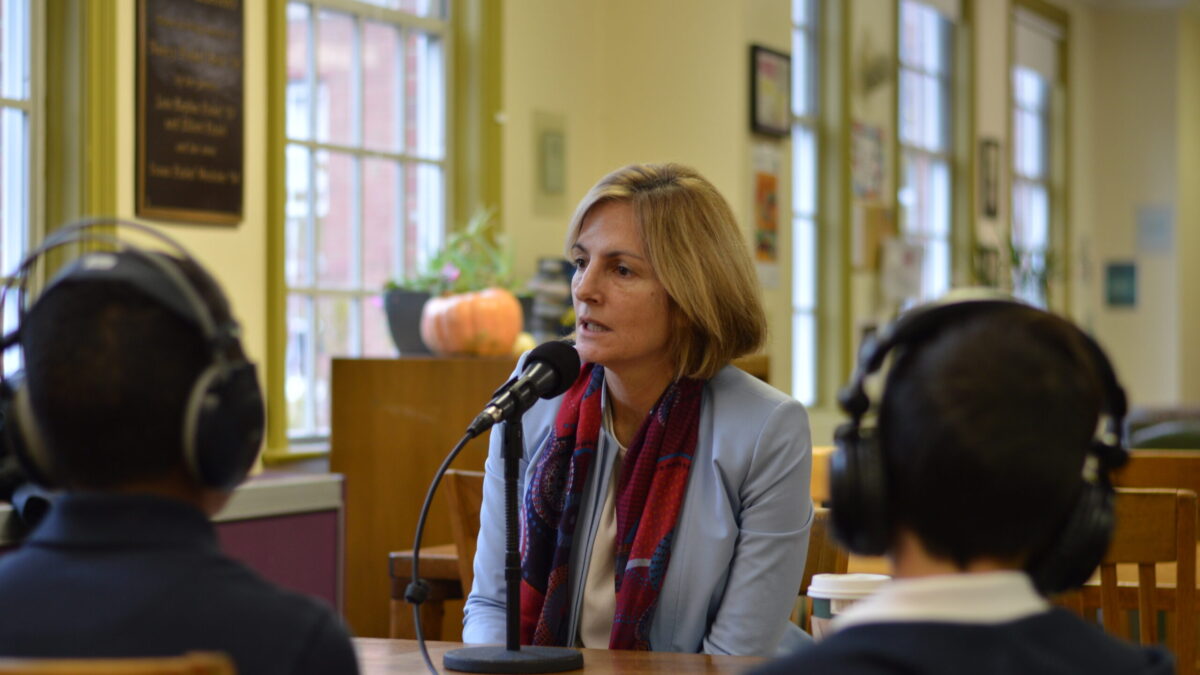 Two youth wearing headphones interviewing an adult speaking into a microphone on a wooden table