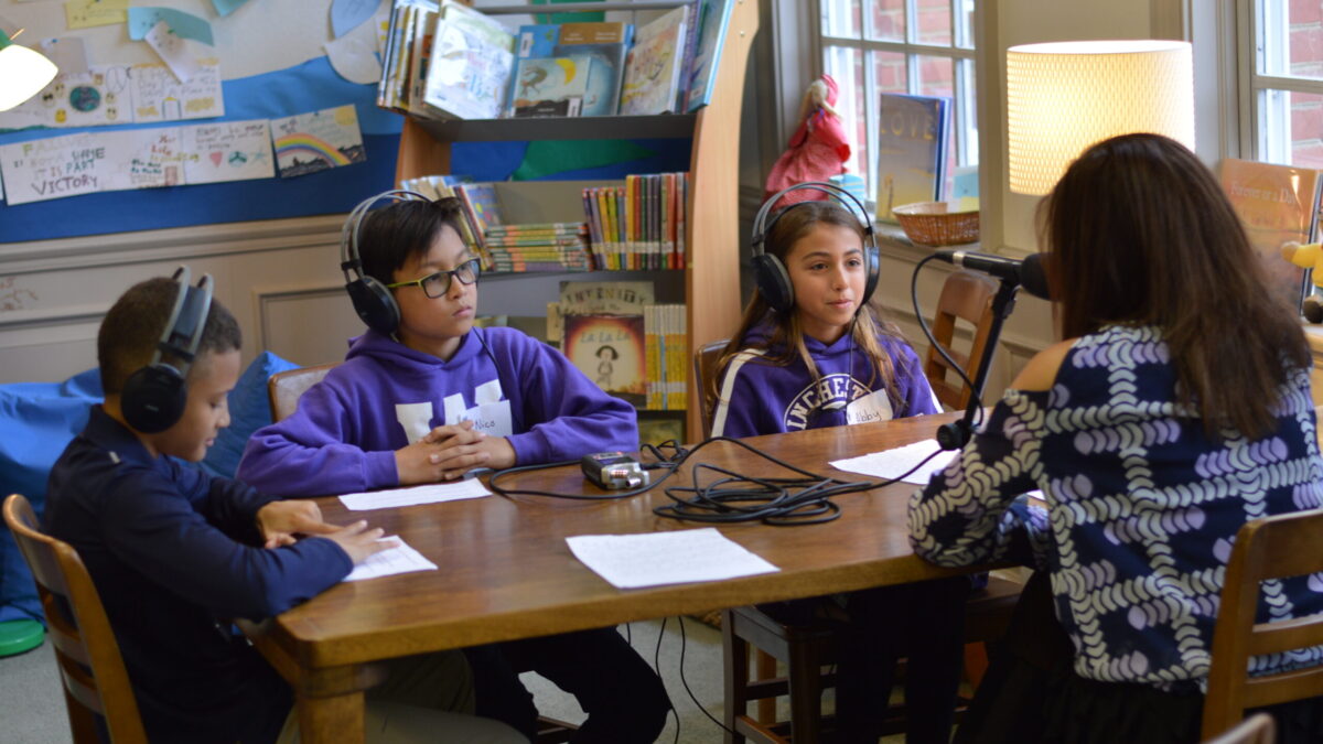 Three youth wearing headphones interviewing an adult speaking into a microphone connected to a portable recorder on a wooden table in a classroom