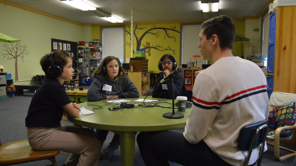 Three youth wearing headphones interviewing an adult speaking into a microphone connected to a portable recorder in a classroom
