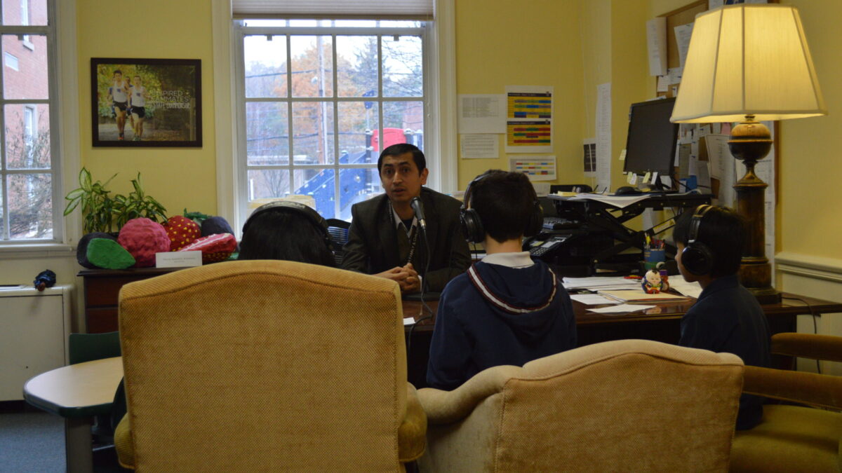 Three youth wearing headphones interviewing an adult speaking into a microphone on a wooden desk in a office