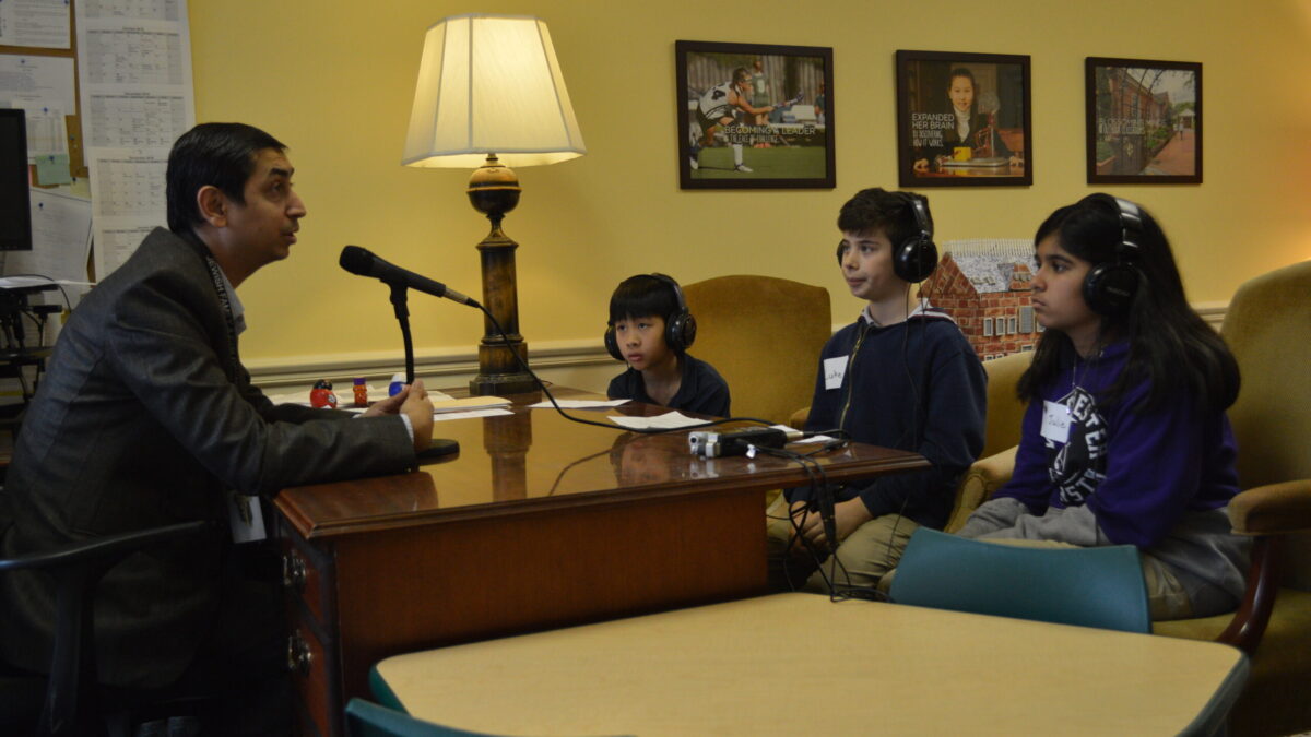Three youth wearing headphones interviewing an adult speaking into a microphone connected to a portable recorder on a wooden desk