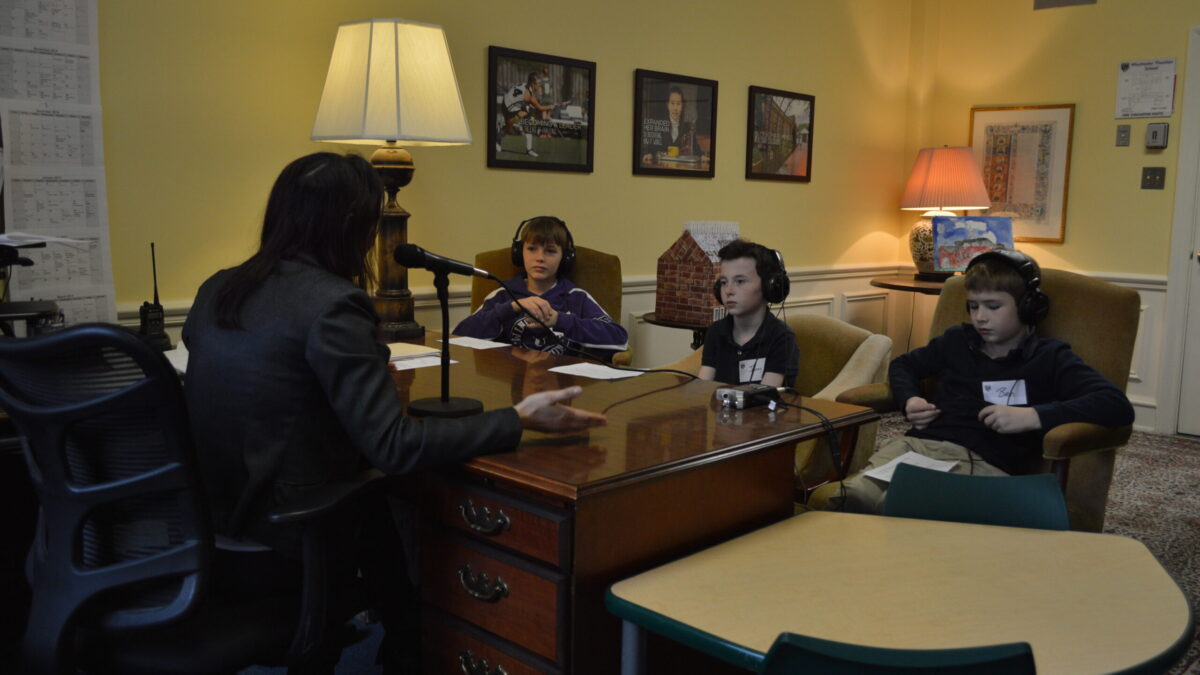 Three youth wearing headphones interviewing an adult speaking into a microphone connected to a portable recorder on a wooden desk in an office