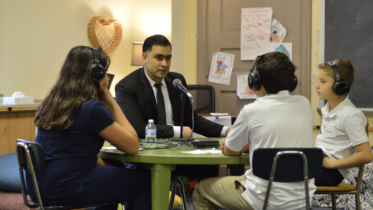 Three youth wearing headphones interviewing an adult speaking into a microphone connected to a portable recorder on a green table in a classroom