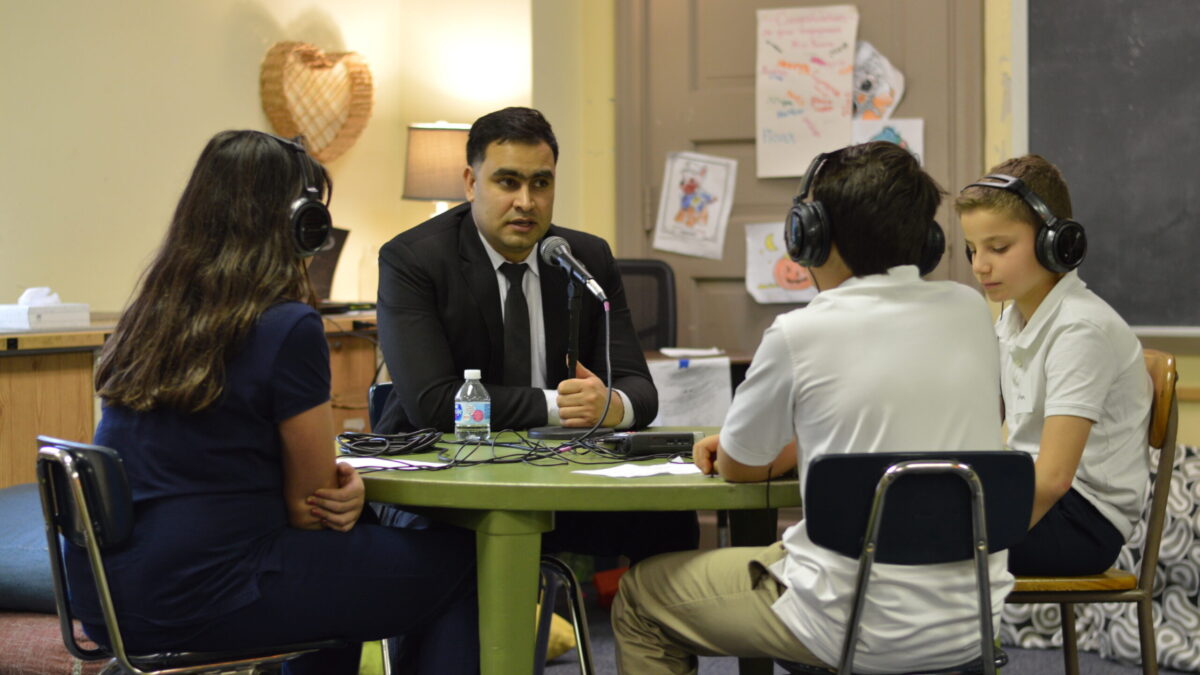 Three youth wearing headphones interviewing an adult speaking into a microphone connected to a portable recorder on a green table in a classroom