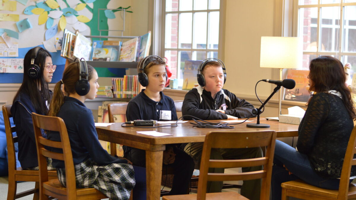 Four youth wearing headphones interviewing an adult speaking into a microphone connected to a portable recorder on a wooden table in a classroom