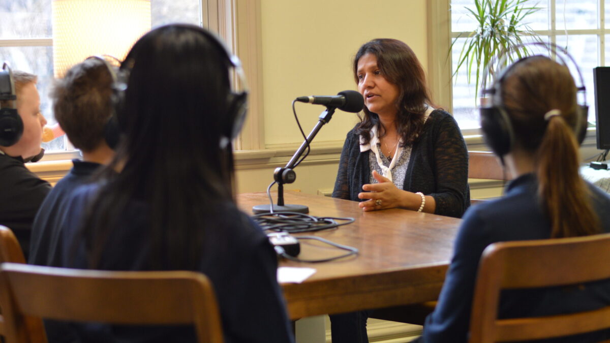 Four youth wearing headphones interviewing an adult speaking into a microphone connected to a portable recorder on a wooden table