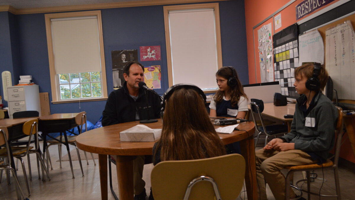 Three youth wearing headphones interviewing an adult speaking into a microphone connected to a portable recorder in a classroom