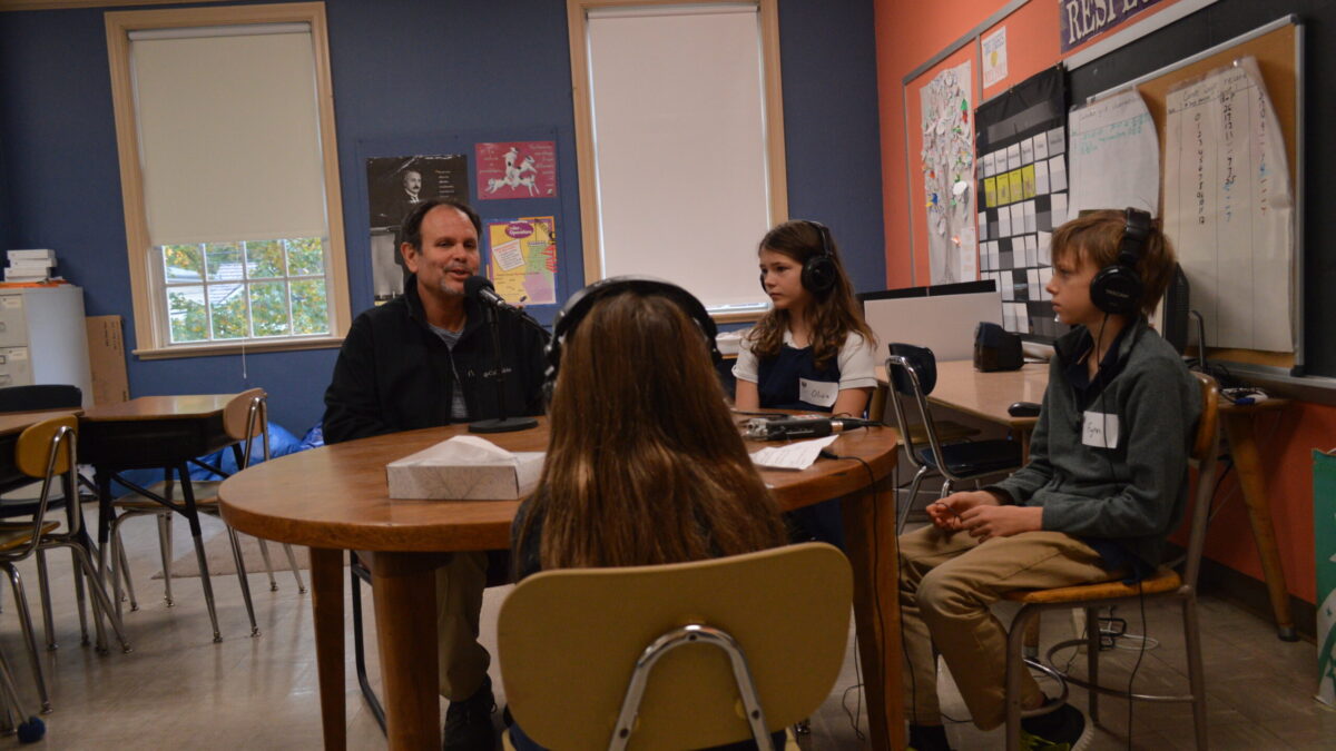 Three youth wearing headphones interviewing an adult speaking into a microphone connected to a portable recorder