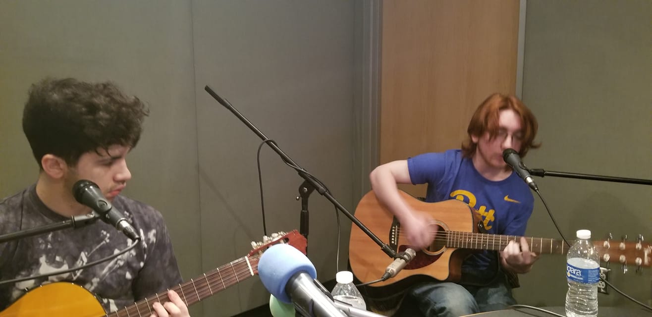Two teens playing guitar and singing into microphones in a radio studio
