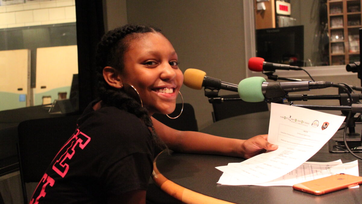 A teen sitting behind a microphone holding a piece of paper and smiling in profile in a radio studio