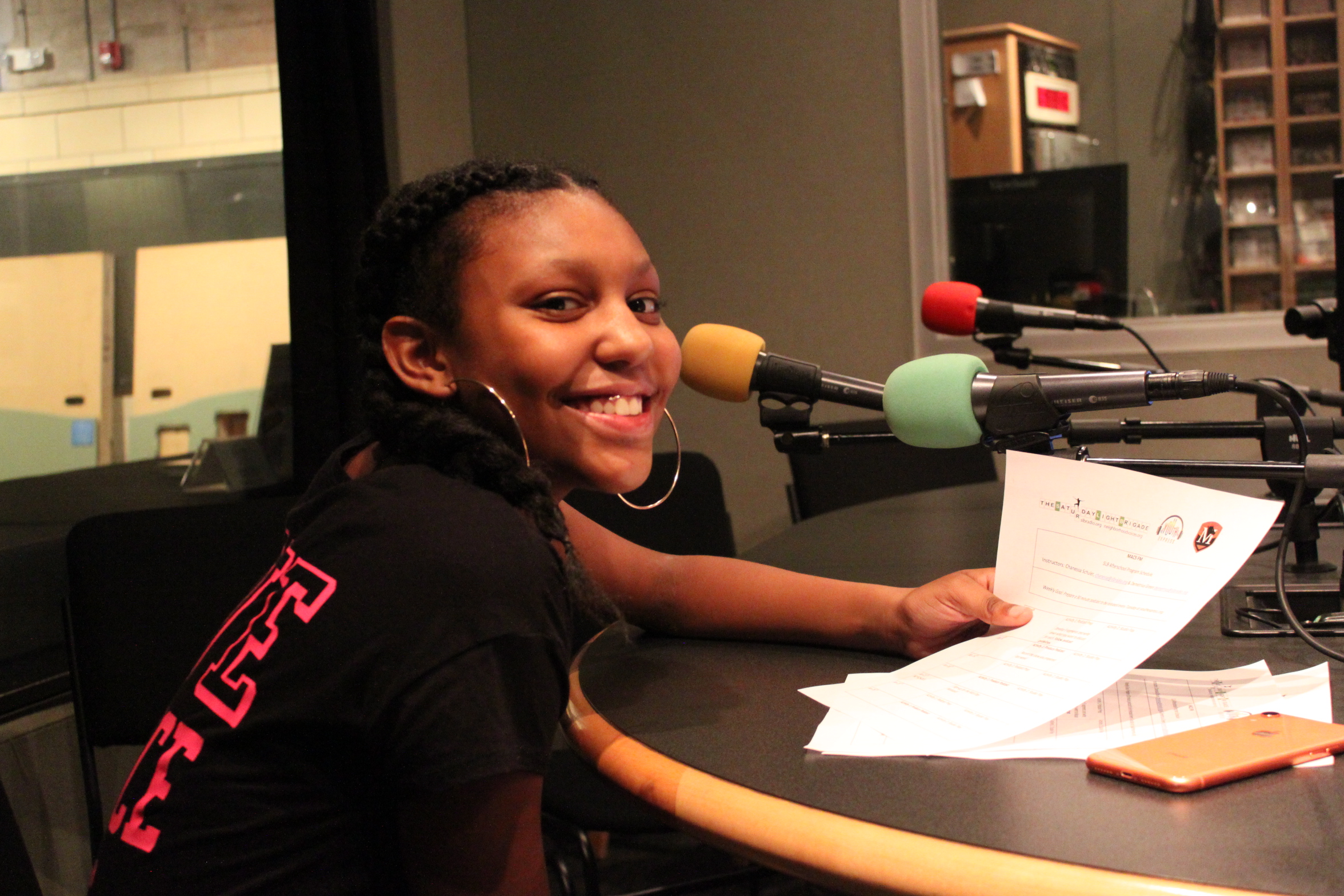 A teen sitting behind a microphone holding a piece of paper and smiling in profile in a radio studio