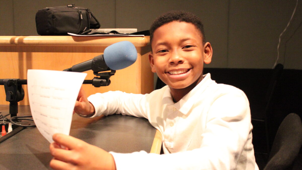 Child sitting in front of a microphone holding a piece of paper and smiling in profile in a radio studio