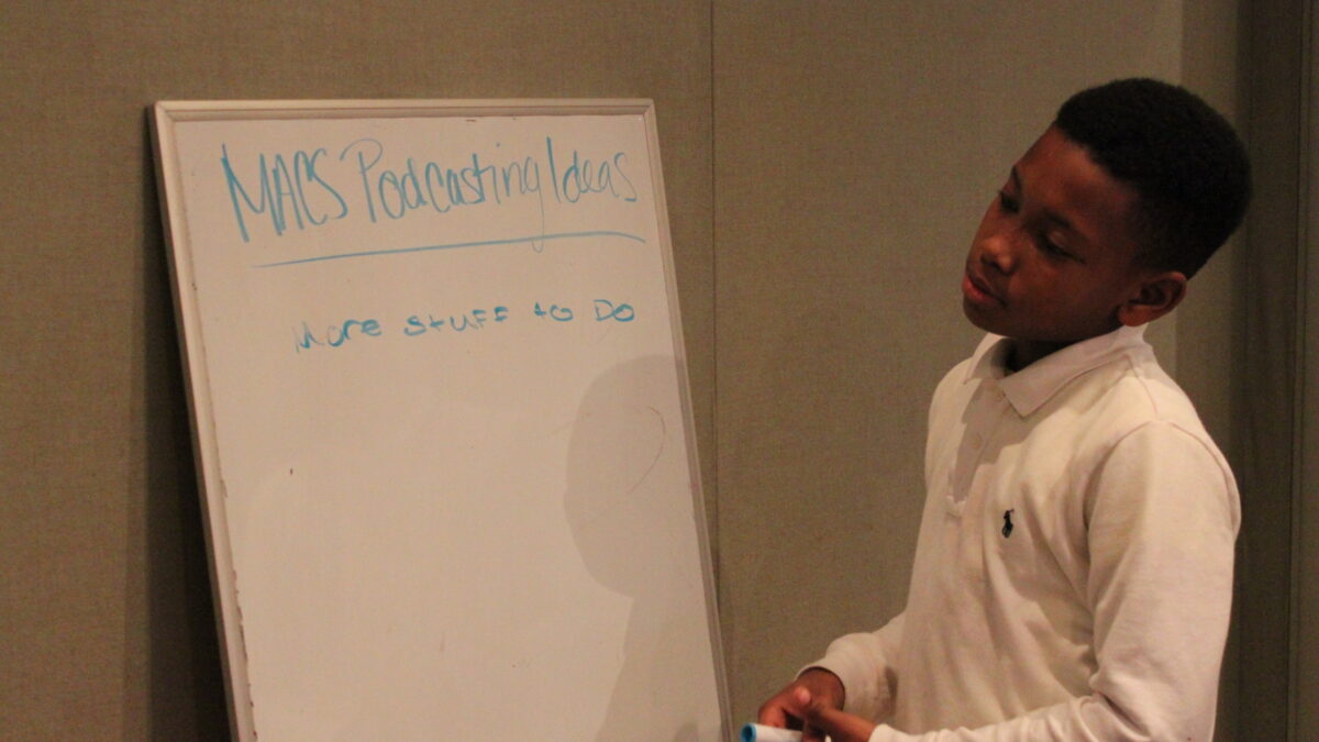 A youth holding light blue markers looking at a white board in a radio studio