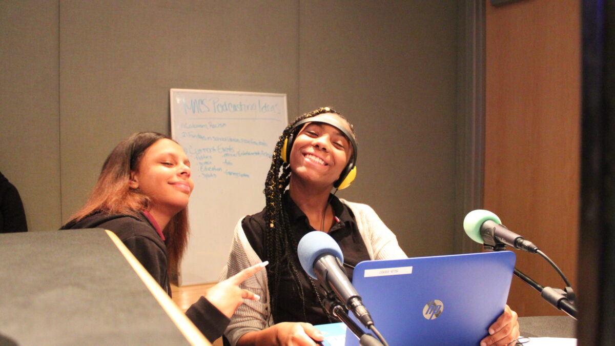 Two teens, one wearing headphones, sitting behind microphones looking at a blue laptop in a radio studio