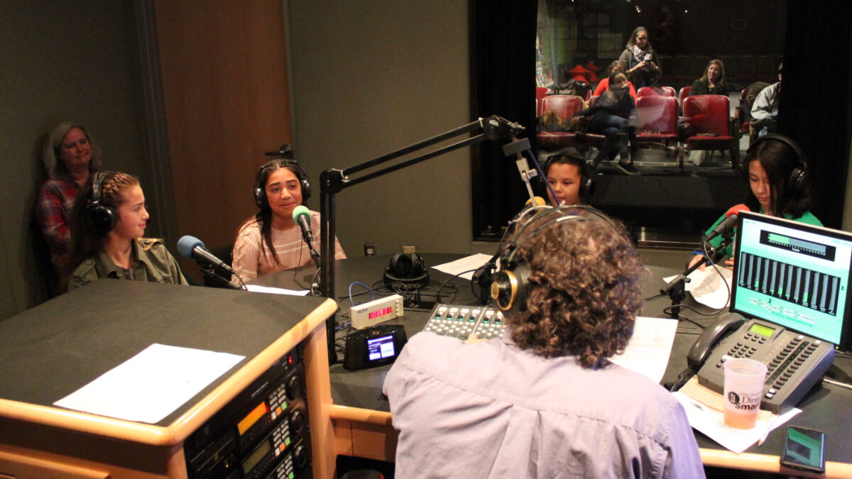 Four youth wearing headphones speaking into microphones being interviewed by an adult wearing headphones sitting behind a microphone and a mixing board as another adult supervises in the background in a radio studio