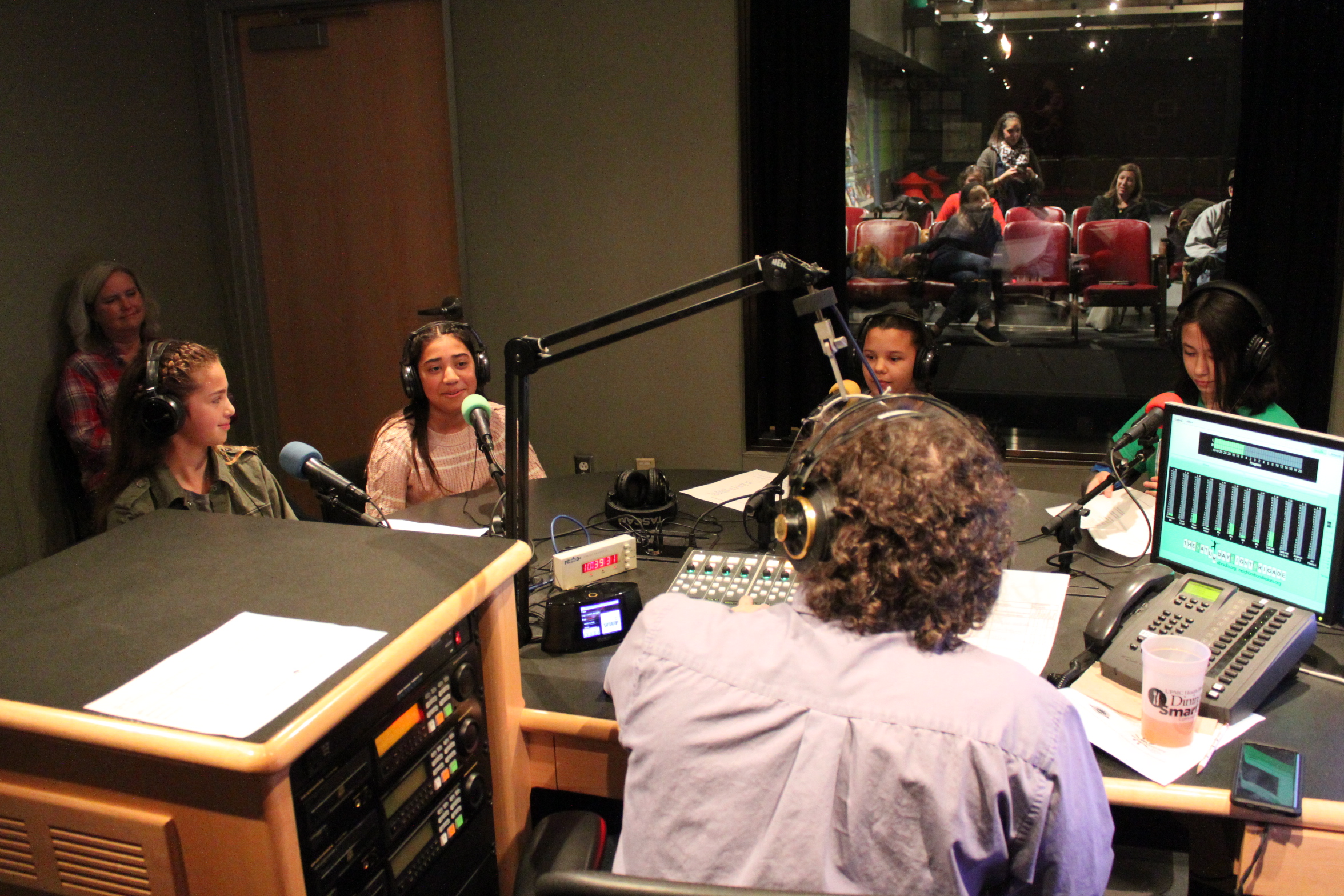 Four youth wearing headphones speaking into microphones being interviewed by an adult wearing headphones sitting behind a microphone and a mixing board as another adult supervises in the background in a radio studio
