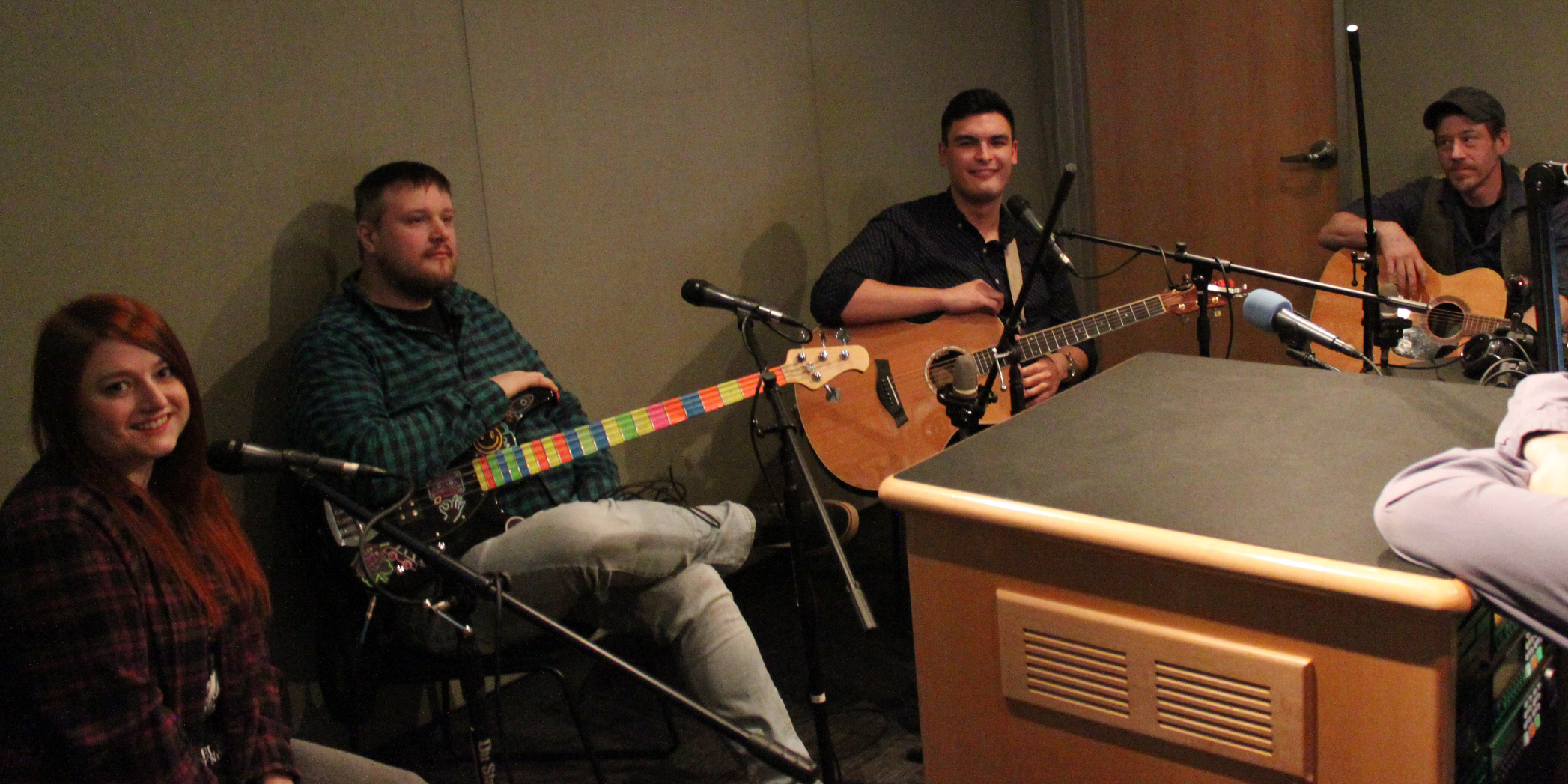 Four adults sitting behind microphones, two holding guitars and one holding a bass, in a radio studio