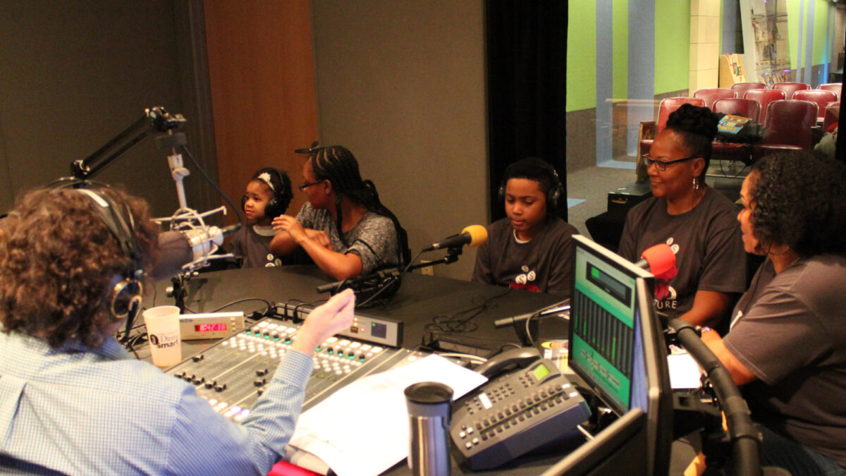 Three adults and two children wearing headphones being interviewed by an adult wearing headphones speaking into a microphone sitting behind a mixing board and gesturing with their right hand in a radio studio