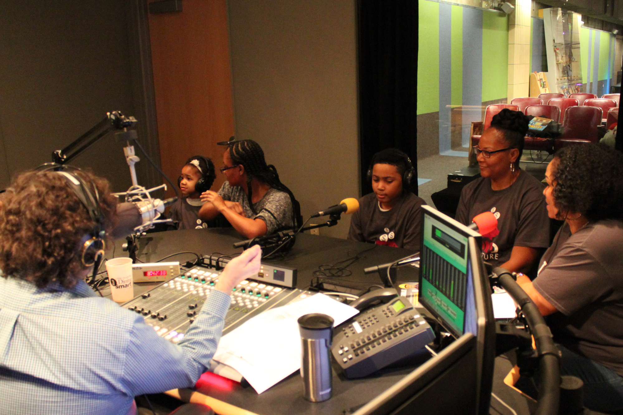 Three adults and two children wearing headphones being interviewed by an adult wearing headphones speaking into a microphone sitting behind a mixing board and gesturing with their right hand in a radio studio