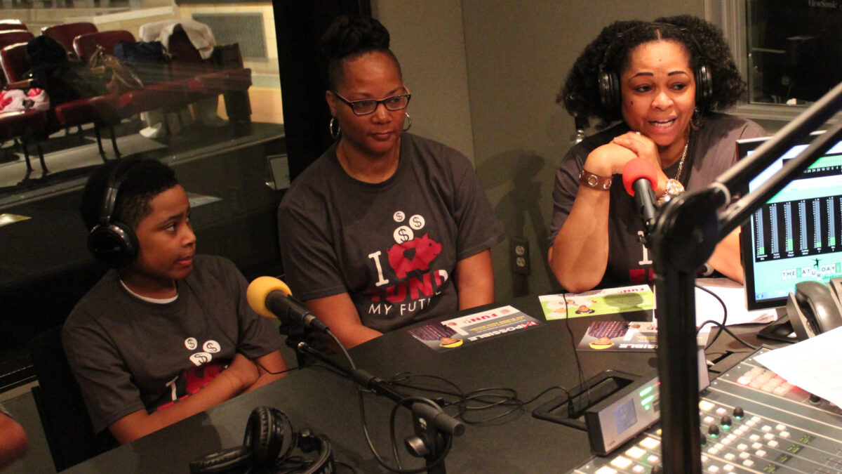 An adult wearing headphones speaking into a microphone next to another adult and a child wearing headphones in a radio sutdio
