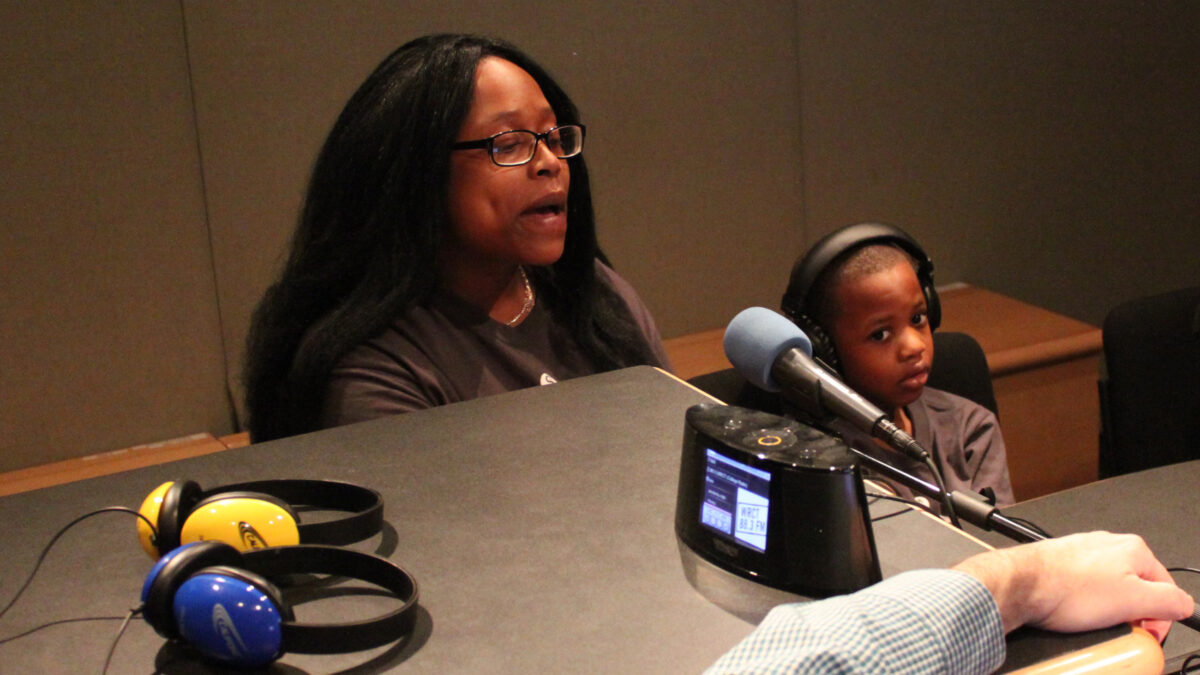 An adult speaking into a microphone next to a child wearing headphones in a radio studio