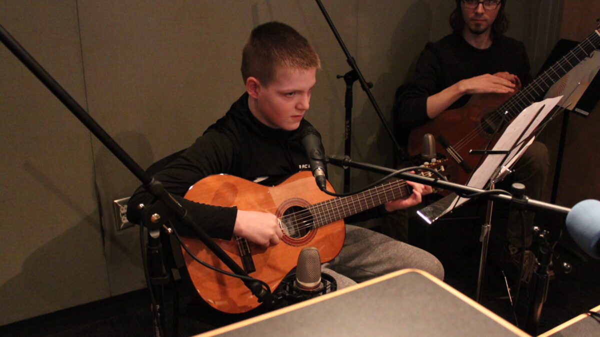 An adult and a youth playing guitar looking at sheet music on a music stand in a radio studio