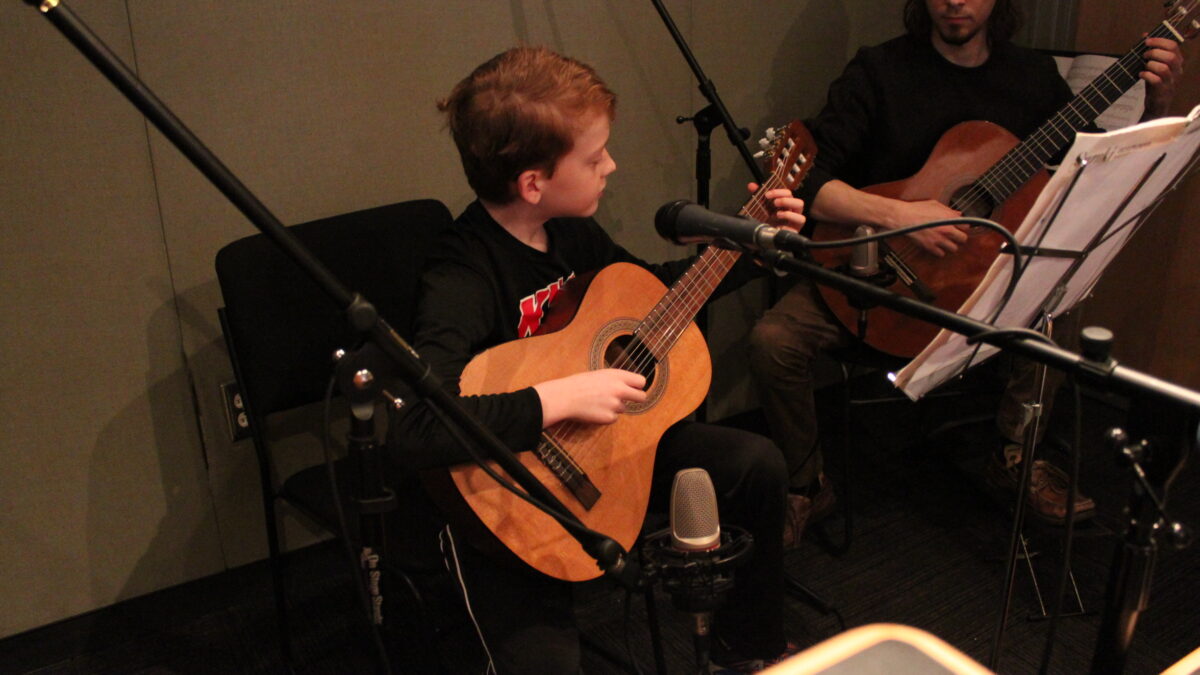 A child and an adult playing guitar and looking at sheet music on a music stand in a radio studio