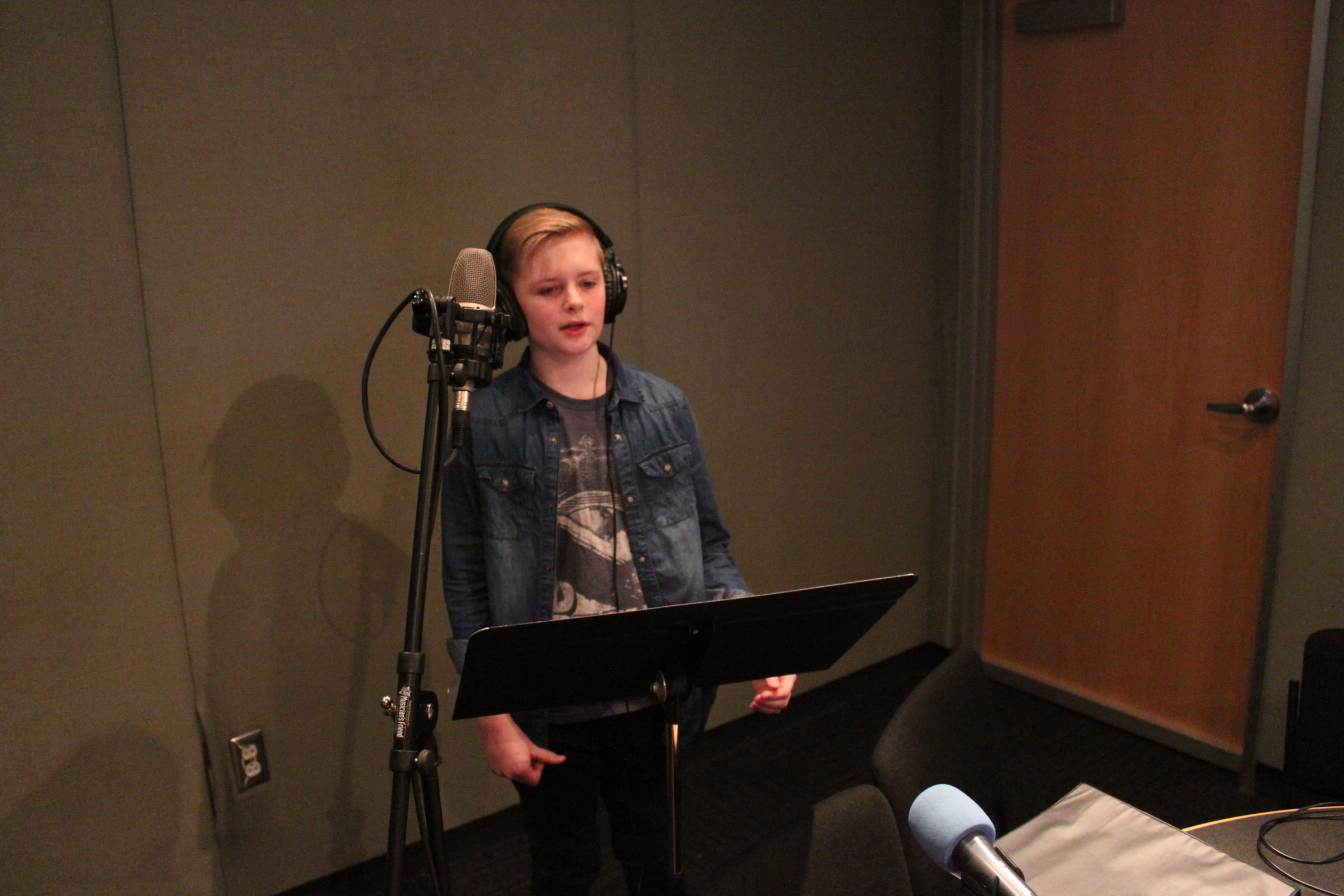 A child wearing headphones singing into a microphone in front of a music stand in a radio studio
