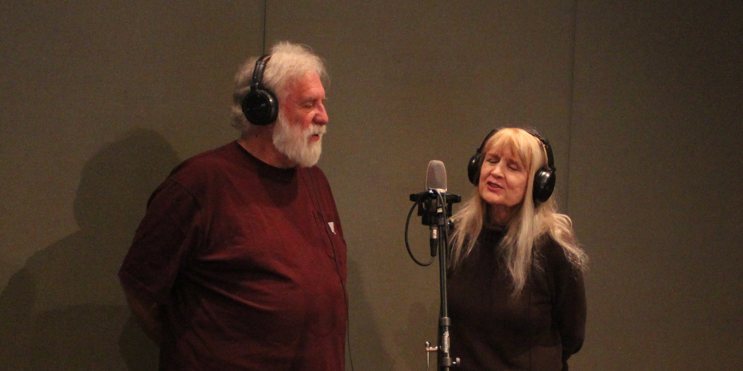Two adults wearing headphones singing into a microphone in a radio studio