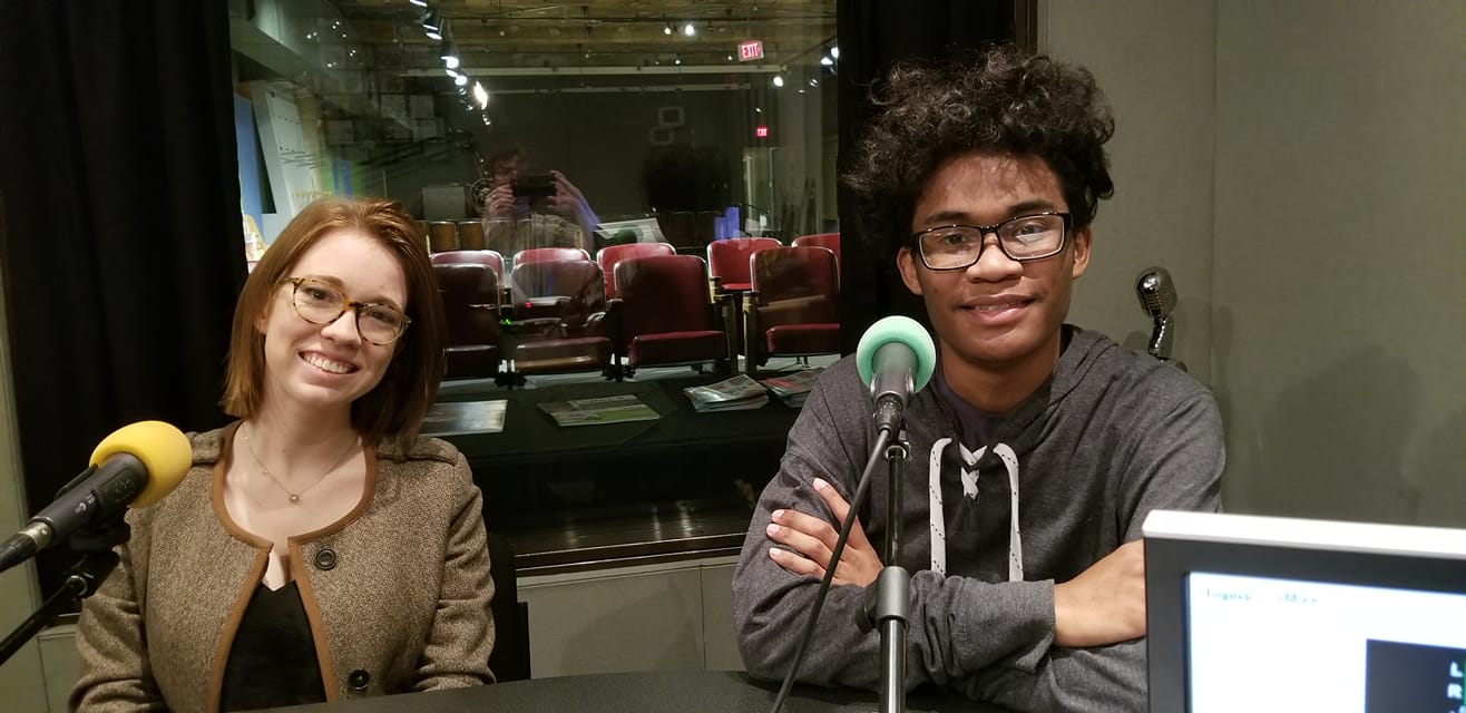 Two teens wearing glasses sitting behind microphones in a radio studio