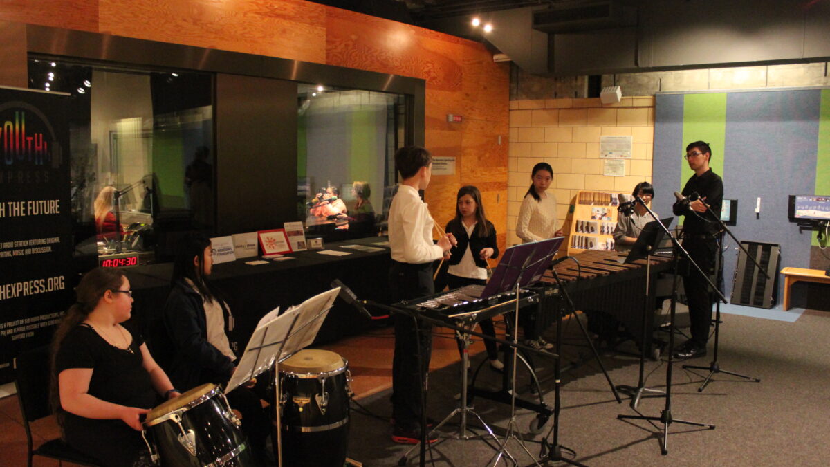 Six youth sitting behind congas, a xylophone, a marimba, and a piano keyboard as an adult looks on