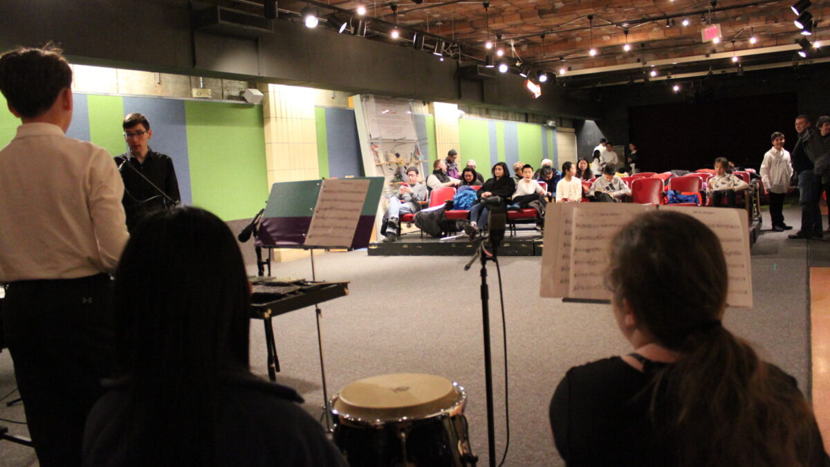 Three youth sitting behind congas and a xylophone looking at an adult in front of an audience of adults