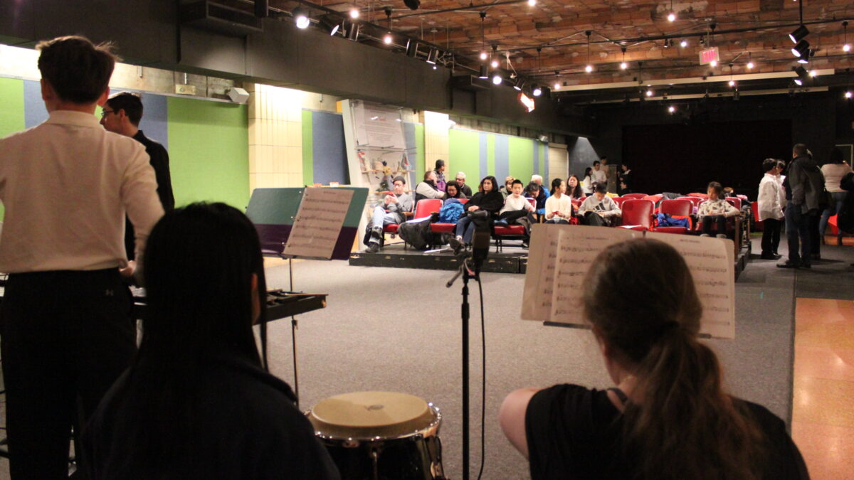Three youth sitting behind congas and a xylophone looking at an adult in front of an audience of adults