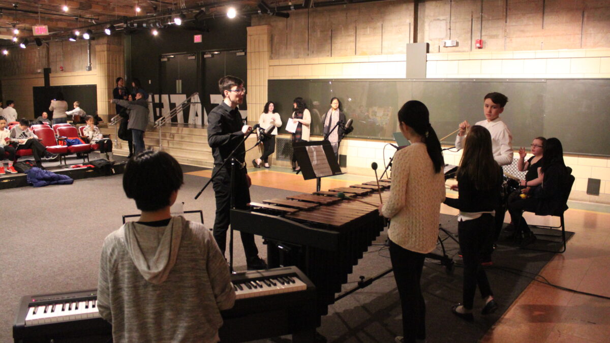 An adult conducting youth performing on congas, xylophone, marimba, and piano keyboard in front of an audience of adults