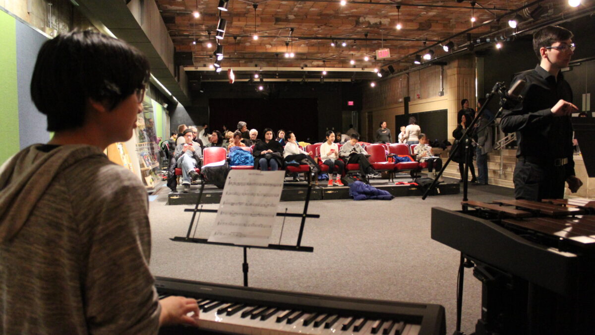 Youth playing piano keyboard looking at an adult conducting in front of an audience of adults