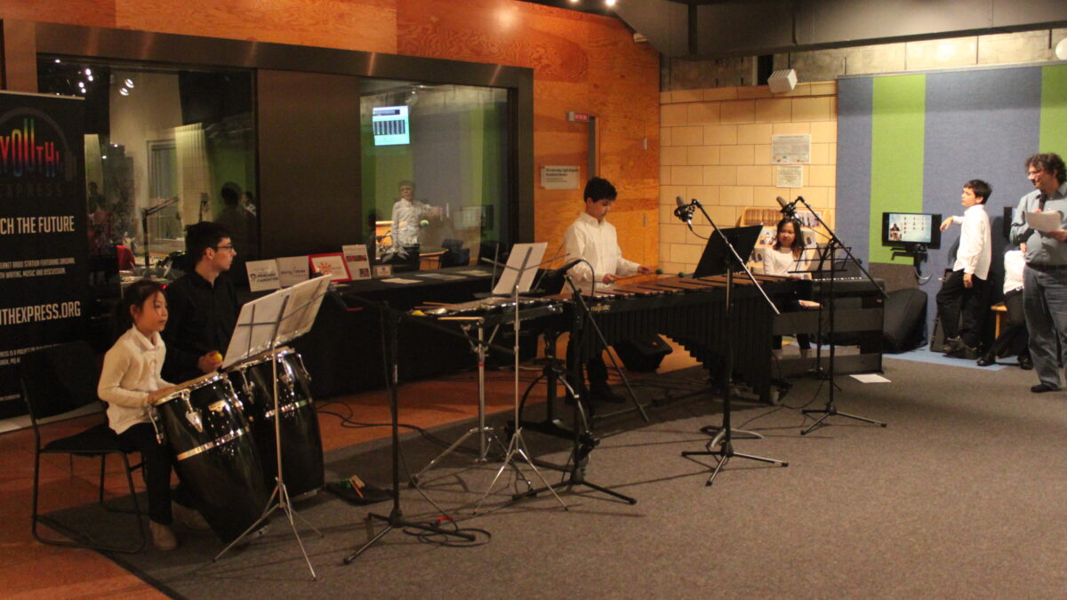 Four youth performing on congas, marimba, and piano keyboard looking at sheet music on music stands