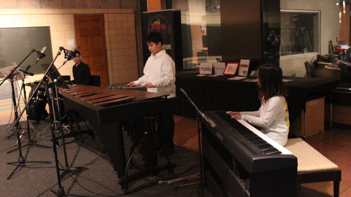 Three youth playing congas, marimba, and piano keyboard looking at sheet music on music stands