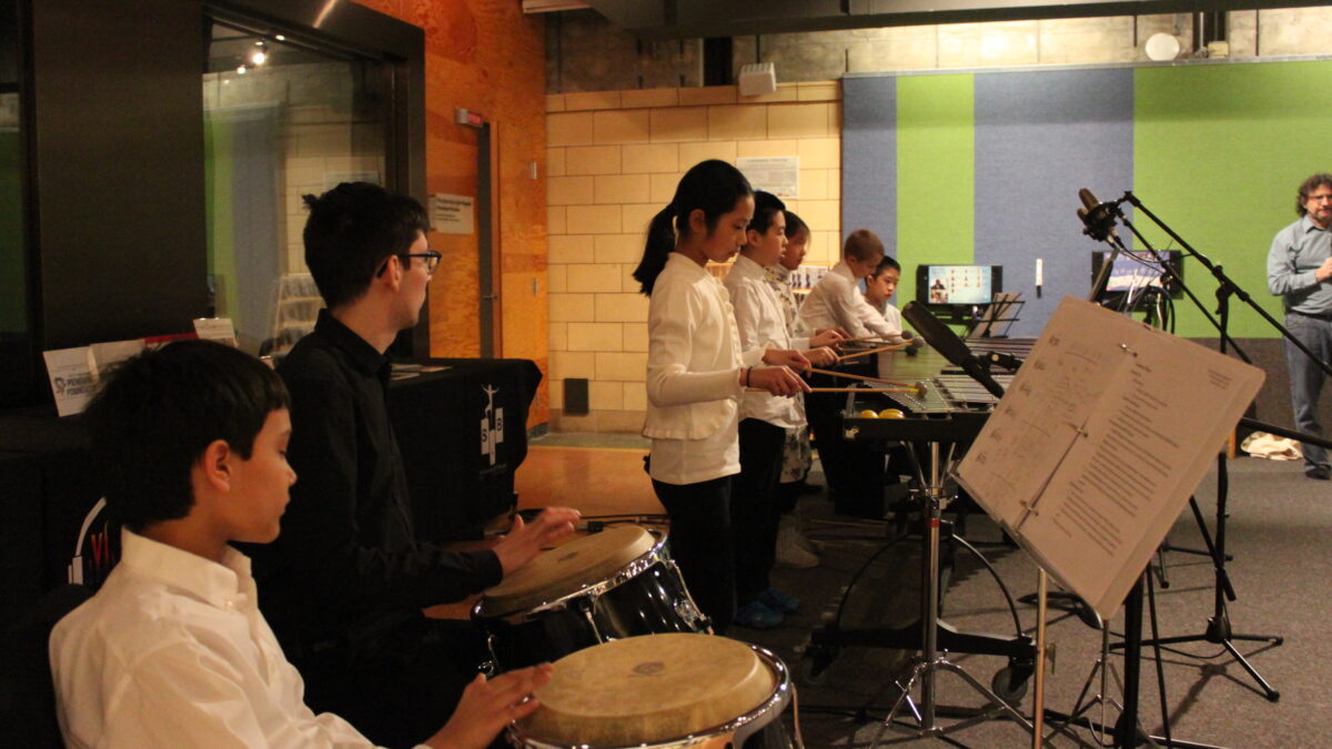 Seven youth performing on congas, xylophone, marimba, and piano keyboard looking at sheet music on music stands