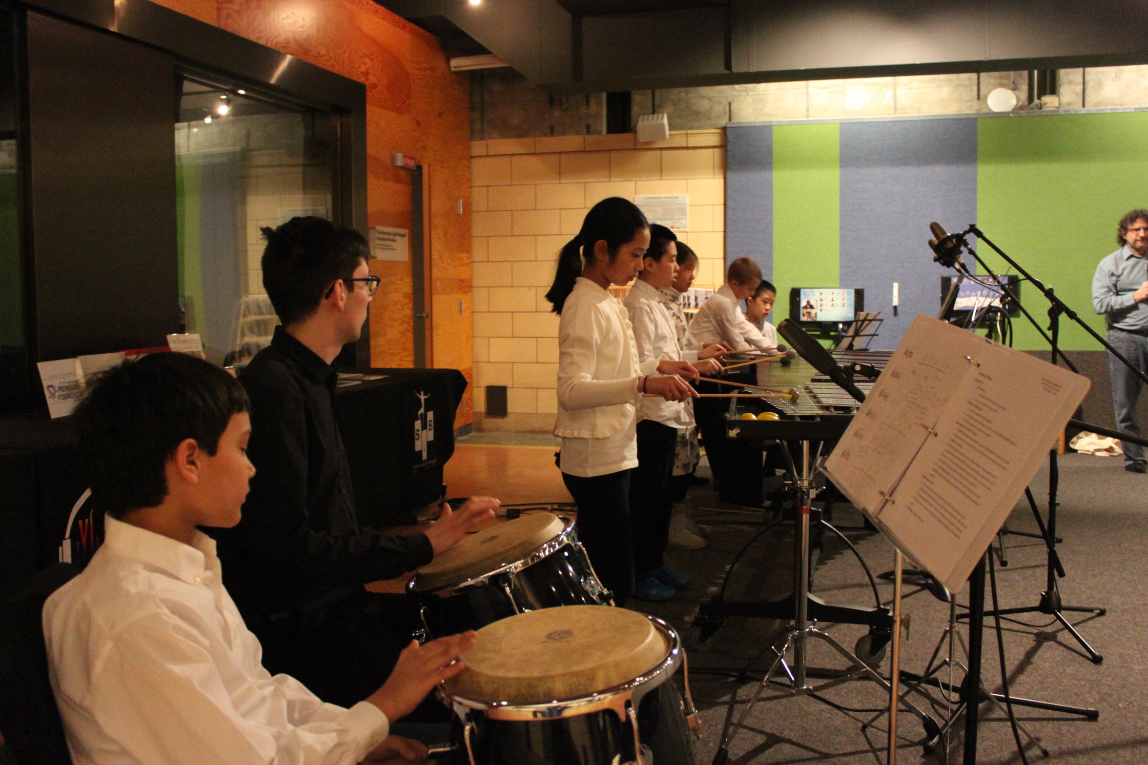 Seven youth performing on congas, xylophone, marimba, and piano keyboard looking at sheet music on music stands