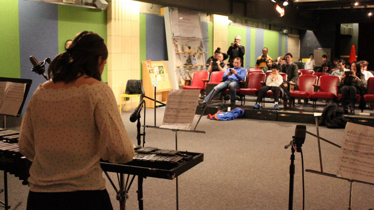 Teen playing xylophone looking at sheet music on a music stand while an audience of adults watches and takes photos