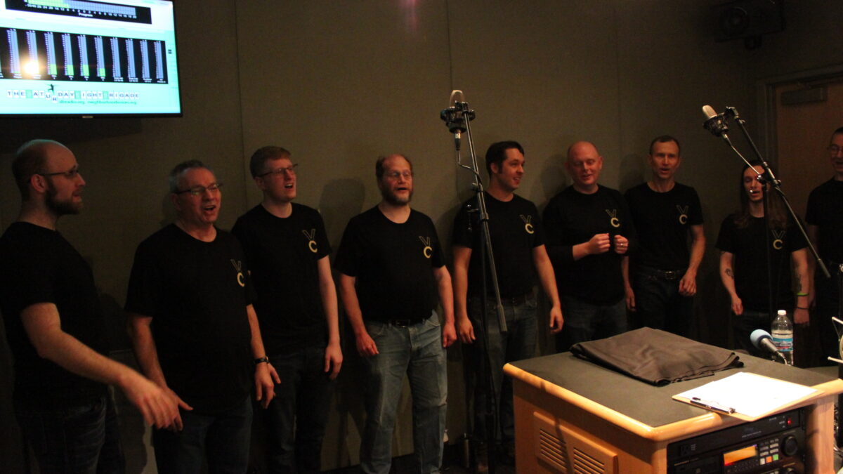 Nine adults singing into microphones in a radio studio