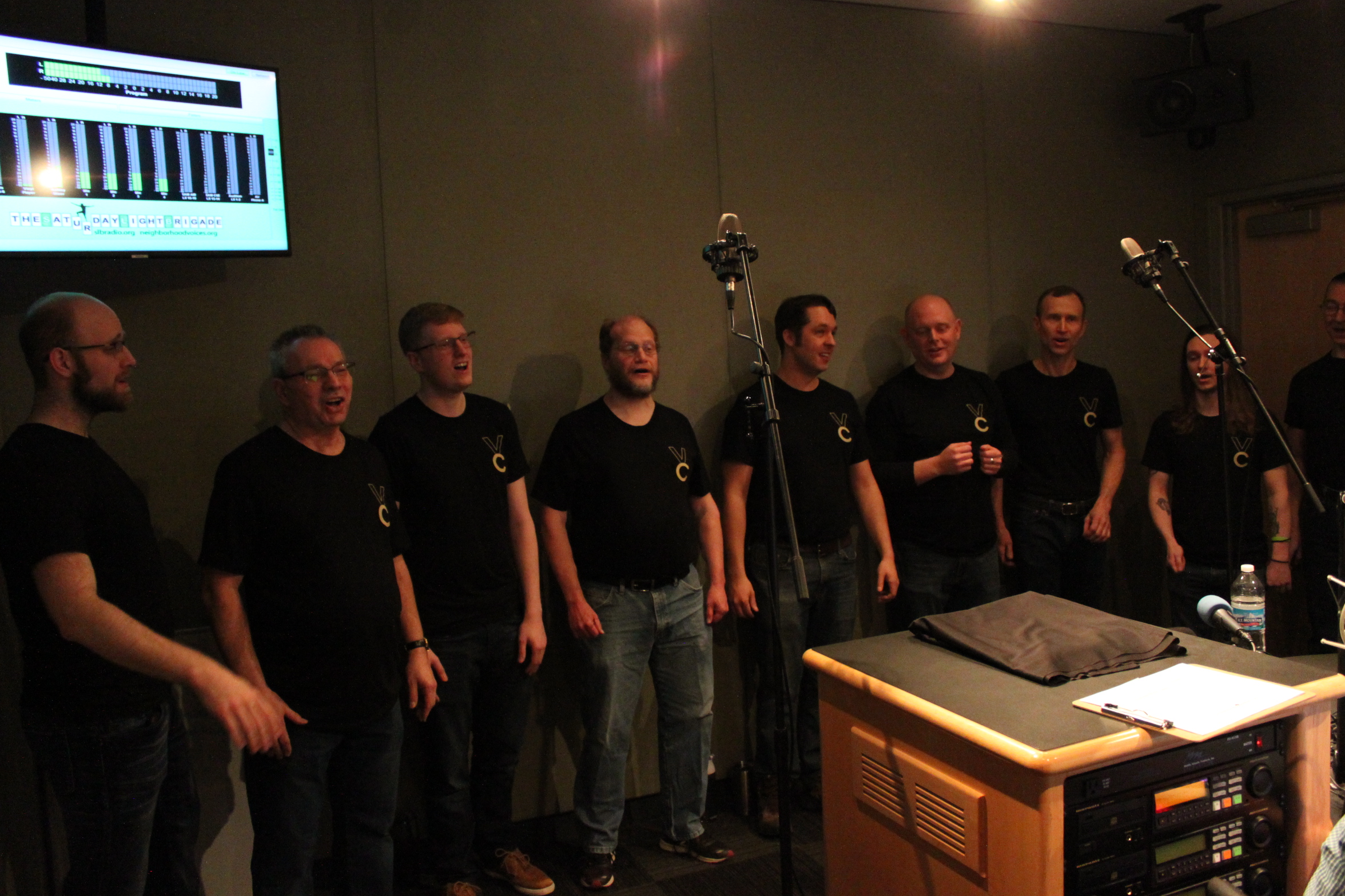 Nine adults singing into microphones in a radio studio