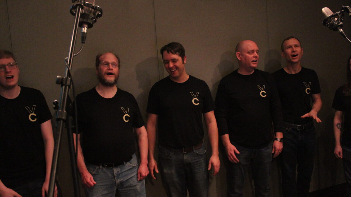 Five adults singing into a microphone in a radio studio