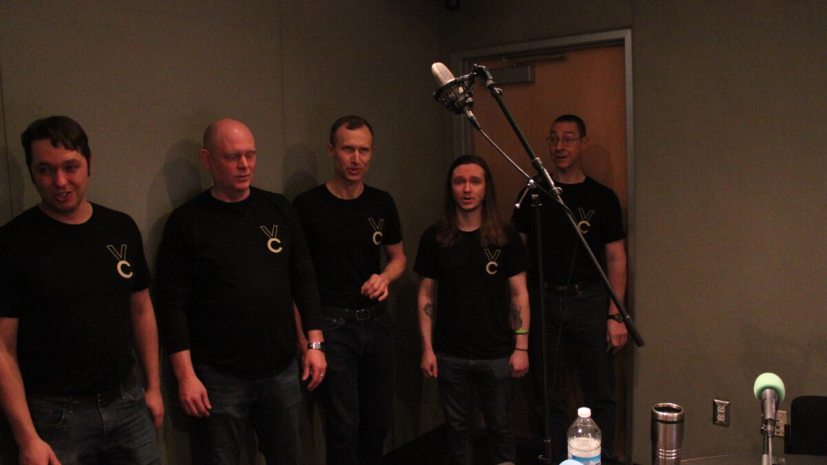 Five adults singing into a microphone in a radio studio