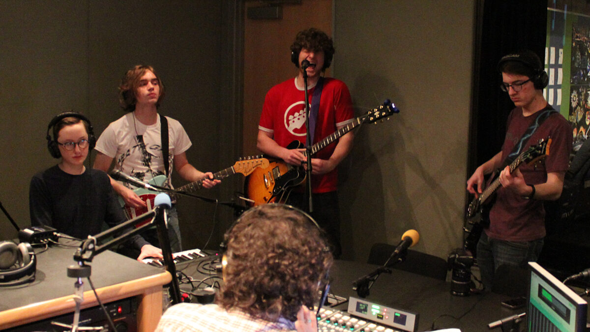 Four teens wearing headphones performing on piano keyboard, guitars, and bass, one of the guitarists singing into a microphone, while an adult listens sitting behind ha mixing board in a radio studio