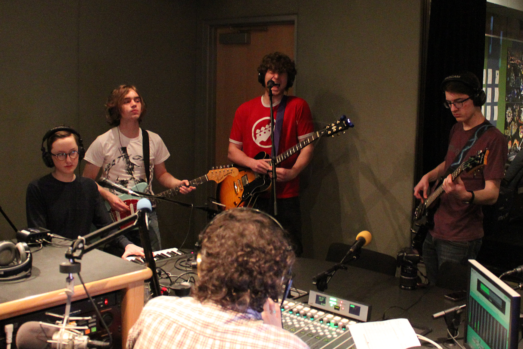 Four teens wearing headphones performing on piano keyboard, guitars, and bass, one of the guitarists singing into a microphone, while an adult listens sitting behind ha mixing board in a radio studio